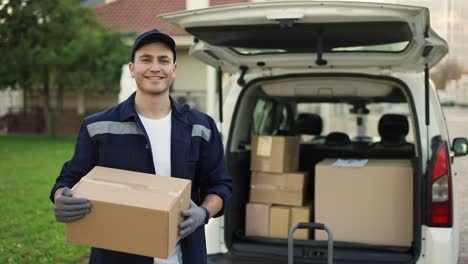 Happy-smiling-delivery-service-worker-in-uniform,-cap-and-gloves-holding-paper-box-and-standing-near-the-minivan-with-opened