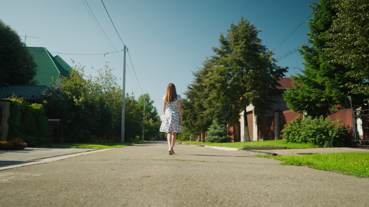 Rear view of confident woman in floral dress walking alone down quiet suburban street while swinging arms playfully under clear blue sky surrounded by green trees, homes, and overhead power lines