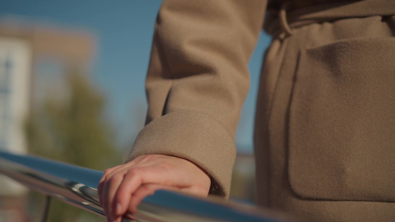 Close up of woman hand resting on and gripping polished metal railings outdoors under clear blue sky. subject wears beige coat sleeves with soft natural light highlighting skin texture