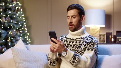 Primer-Plano-Retrato-De-Un-Joven-Feliz-Con-Suéter-De-Navidad-Sentado-En-La-Habitación-Cerca-De-Un-árbol-De-Navidad-Brillante-Decorado-Chateando-En-Línea-Por-Teléfono-Celular