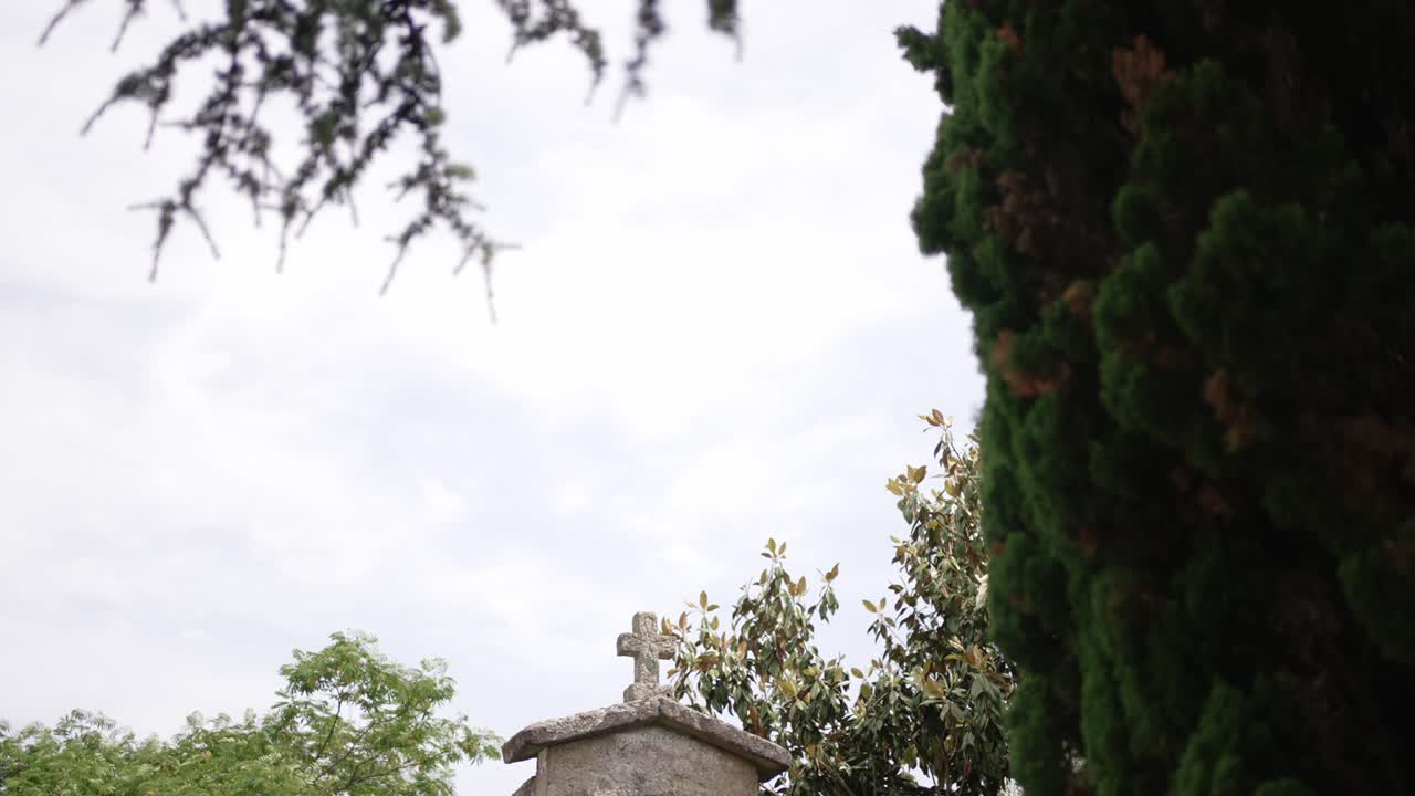 weathered stone cross stands above a small chapel roof framed by green trees and cloudy sky