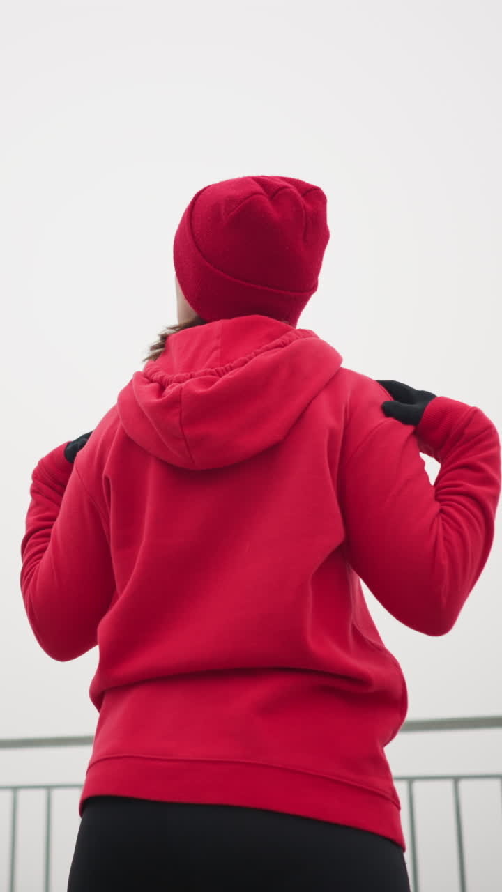 vista trasera de una mujer realizando un estiramiento de la mano girando el brazo y la cabeza hacia la izquierda y la derecha, vestida con ropa atlética, con barandilla de hierro y atmósfera al aire libre serena en el fondo
