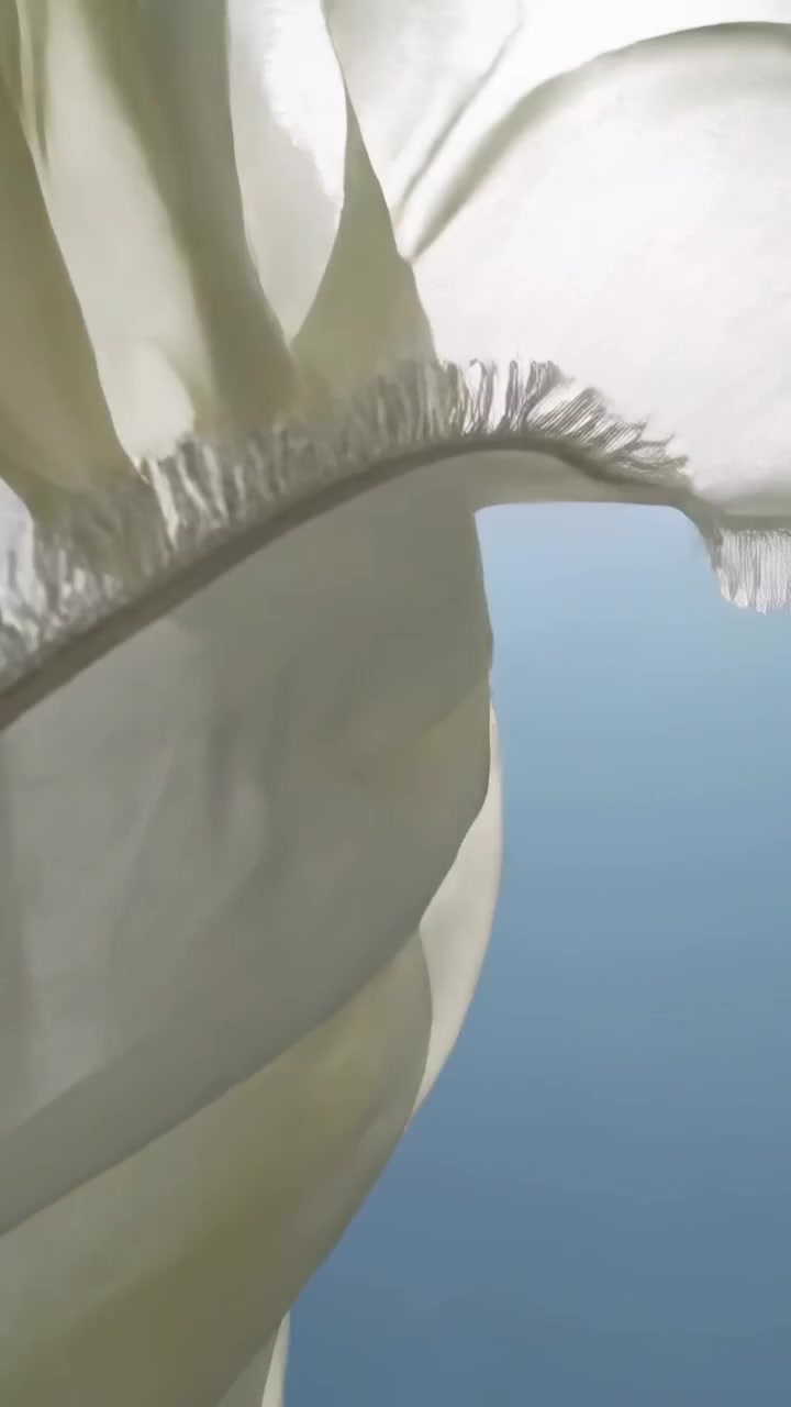 縦型動画：青い空でそよ風により波打ちねじれる膨らむクリーム色の絹、日光を浴びたフリンジと共に