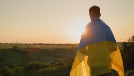 a young man with the flag of ukraine on his shoulders looks at the sunset over a picturesque valley