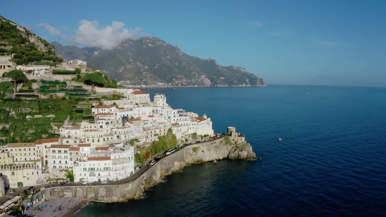 Vista aerea con drone di un'iconica città della Costiera Amalfitana in Italia, con splendidi edifici bianchi a picco sulla scogliera, storico muraglione, spiaggia, profondo Mar Mediterraneo e paesaggio montano.