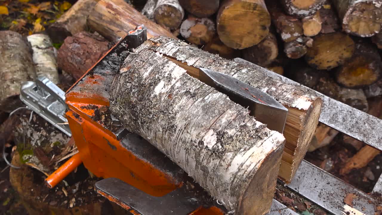 Side view of a birch log timber or firewood material being split in half or into pieces by a wood splitter machine in slow motion at autumn nature with wooden logs in the bokeh blurry background