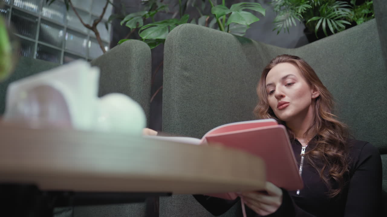 Blonde woman wearing black outfit sits with legs crossed in green booth, holding open pink notebook on lap and thinking deeply, surrounded by indoor plants and blurred tabletop decoration