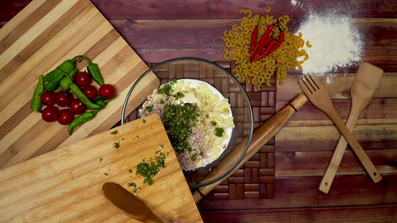 A Top view of adding green chili in to the macaroni, Marinated macaroni in the bowl, raw macaroni, wooden spoon, wooden spatula, flour, rolling pin, a frozen cheese piece and chili on the table