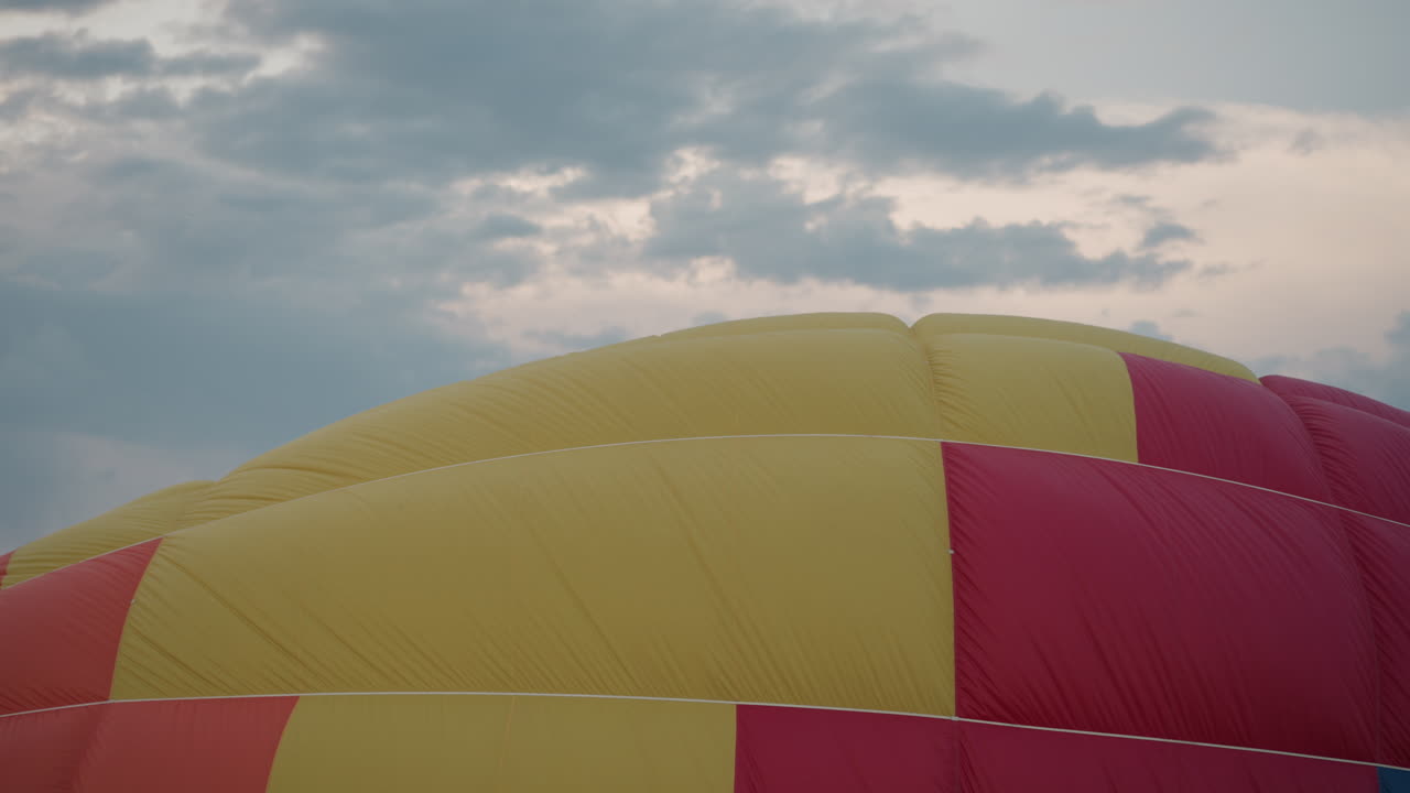 gold and red balloon envelope billowing upward during inflation over grassy field under pastel sky with distant tree line and soft clouds highlighting preflight ascent preparation scene