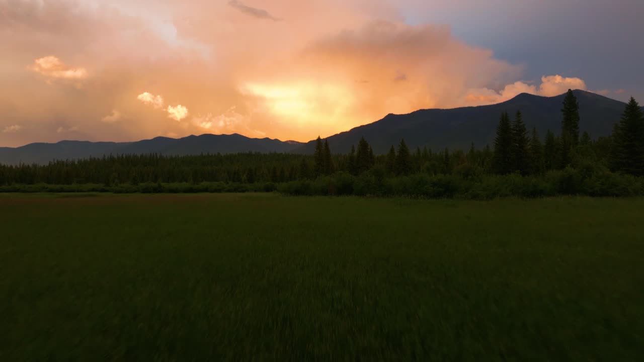 モンタナ山脈の背後にある美しい曇ったオレンジ色の夕日を明らかに緑豊かな緑の谷の上空ドローン飛行