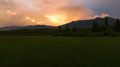 Aerial-drone-fly-over-lush-green-valley-revealing-beautiful-cloudy-orange-sunset-behind-Montana-mountain-range