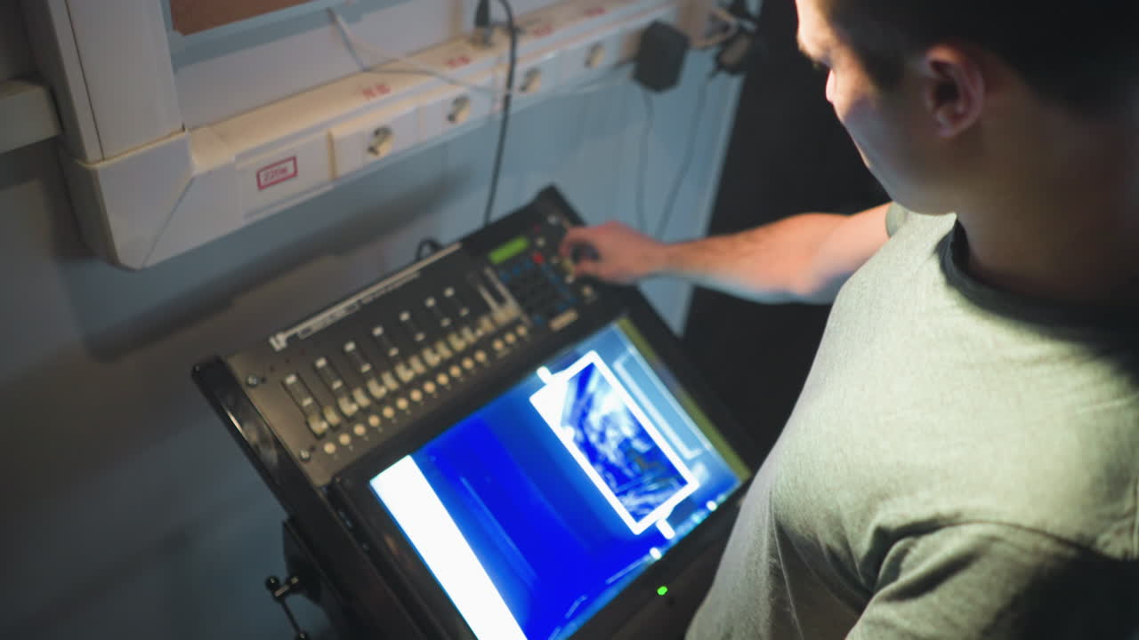 Young man stands focused beside control panel with illuminated touchscreen and fader board in dimly lit studio filled with cables, notes, and technical gear, preparing for operation or stage production task