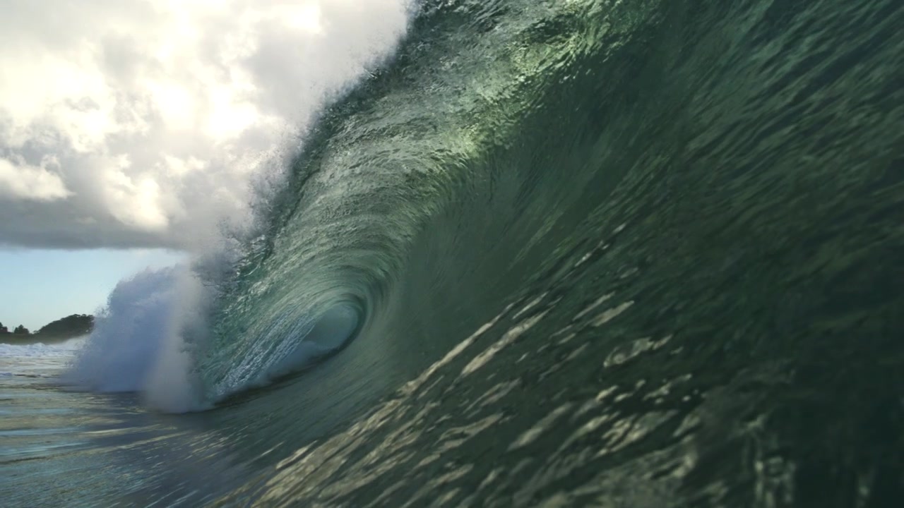 Long green ocean wave rises above reflective water beneath clouds in gentle slow motion