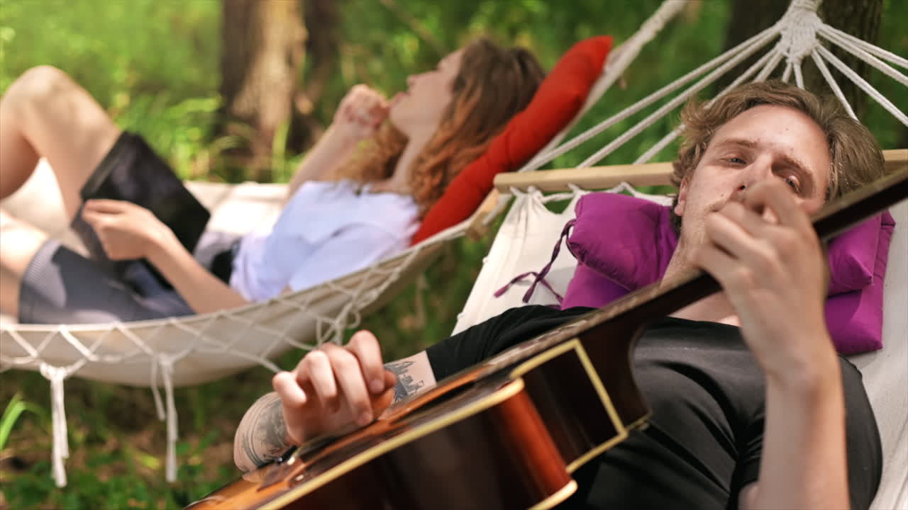Couple lying on hammocks. Young man is playing guitar while woman is using tablet with stylus. Greenery around. Glamping. Slow motion
