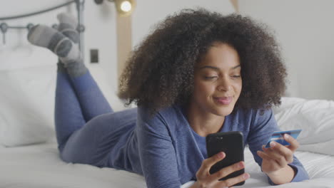 woman lying on bed with mobile phone shopping online using credit card wearing pyjamas