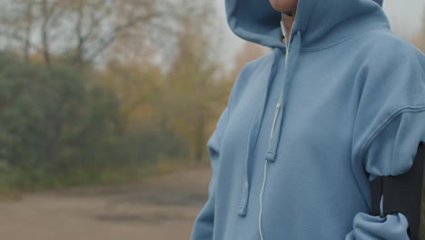 joven deportista seria con capucha azul mirando el reloj inteligente antes de entrenar en el camino del bosque