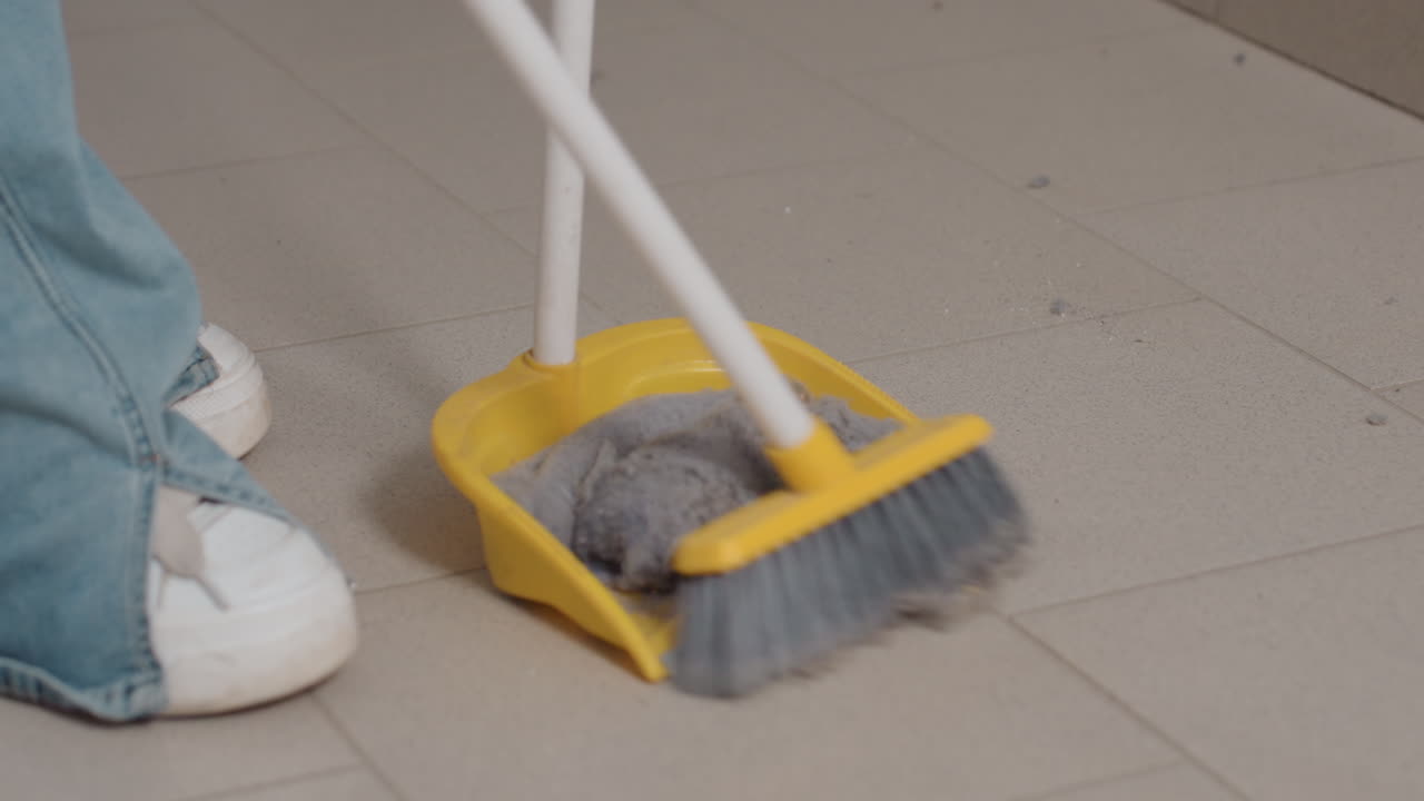 Leg view of staff packing dirt from floor with broom stick and dust pan near industrial washers in laundromat, sweeping lint from filter drawer spill into pan, routine cleanup, maintenance, hygiene