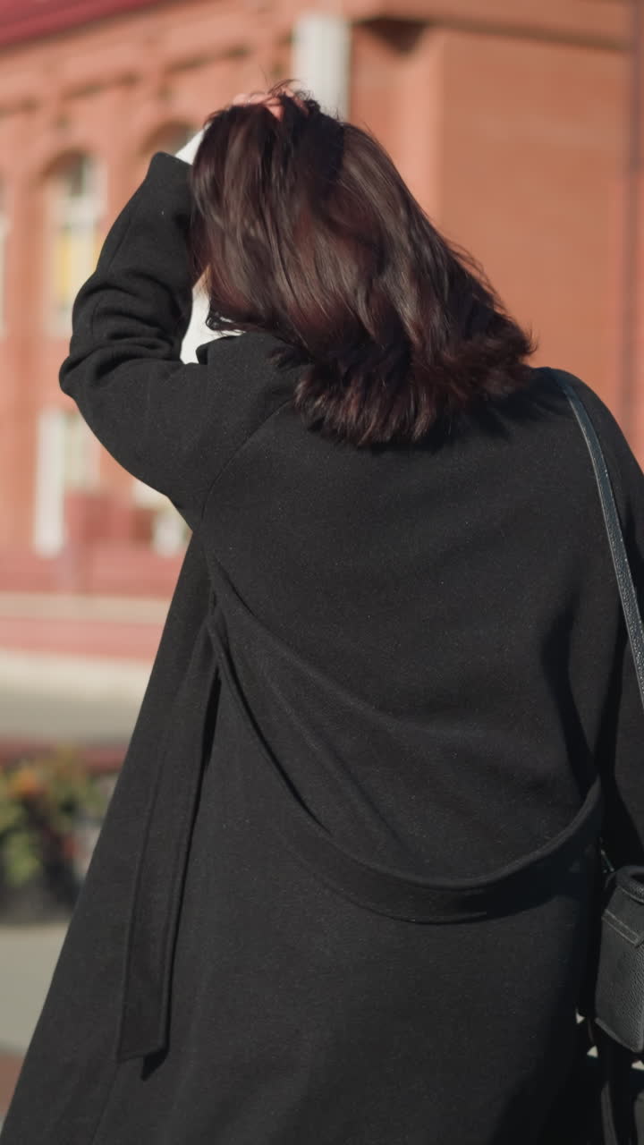 검은 코트와 색 스웨터를 입은 여자가 은 벽돌 건물과 초록색 나무와 함께 은 도시 환경에서 검은 드백을 들고 머리를 뒤로 조정합니다.