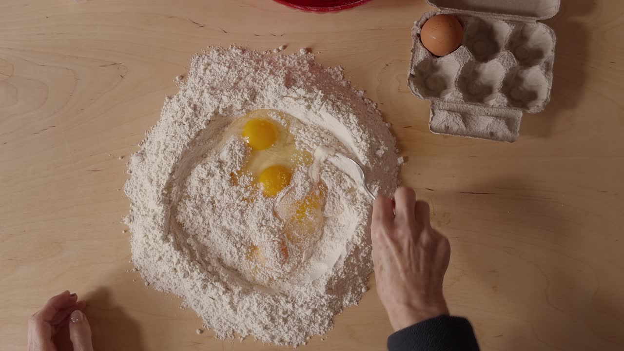 Overhead shot of a Romagna cook mixing eggs and flour with a fork to prepare fresh cappelletti dough. Traditional Italian pasta-making using raw ingredients on a wooden table