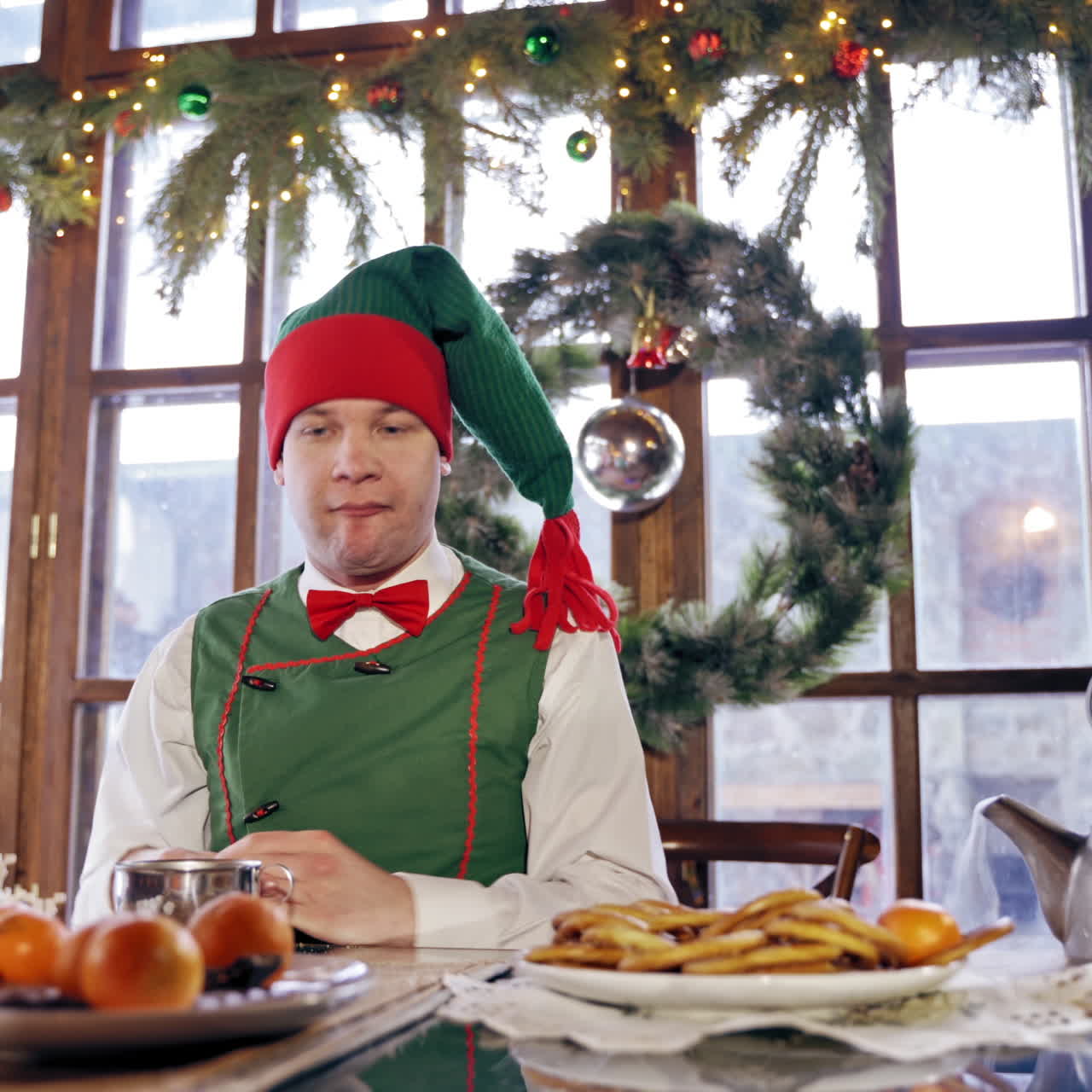 Hungry elf drinking at the table at Christmas. Male elf in green costume with hood eating at festive table on decorated background. New Year and Christmas concept.