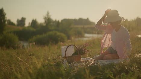 woman in open grassy area under sunlight reaching for sun hat on woven basket filled with flowers, enjoying peaceful summer day outdoors on picnic mat, with blurred natural background