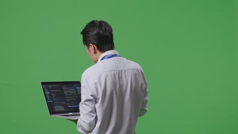 스튜디오에서 녹색 화면 배경에서 노트북 컴퓨터를 사용하는 아시아 사업가의 뒷면, 컴퓨터 디스플레이 코드를 보여줍니다.