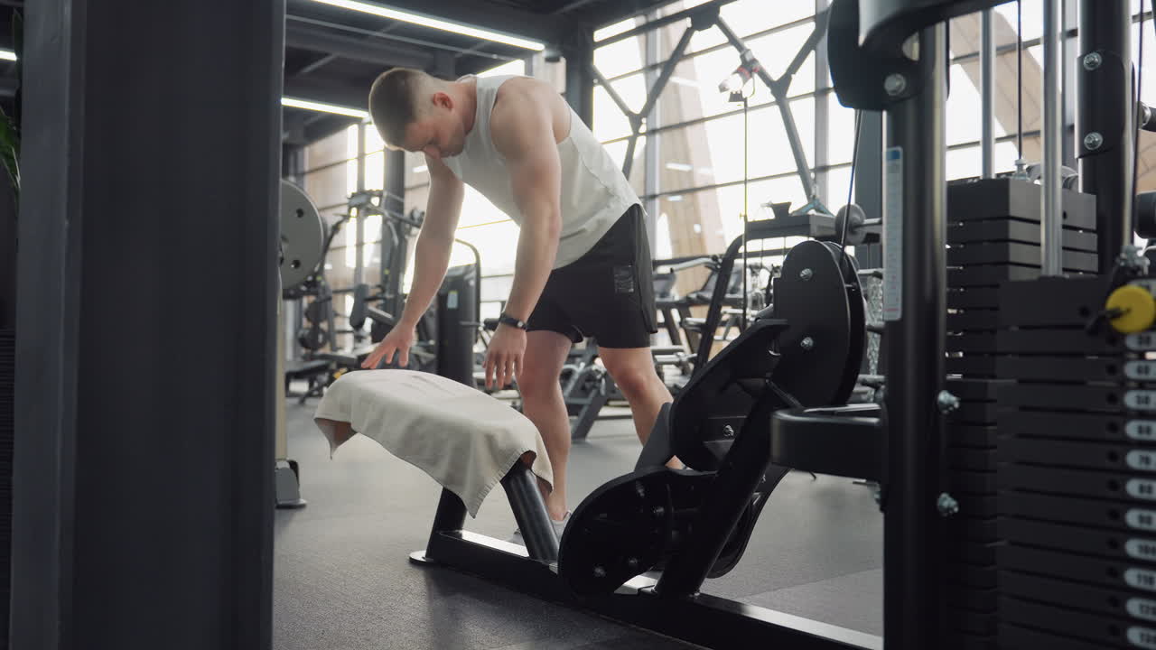 Youth drapes towel over machine bench then reaches to press power switch preparing equipment for next workout in sunlit modern gym environment highlighting hygiene and focused routine momentum