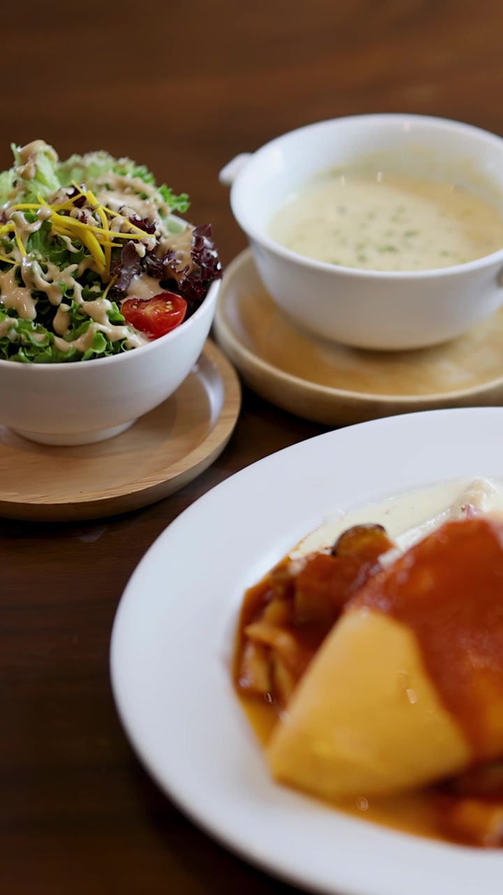 Primer plano cinematográfico en panorámica de ensalada, sopa cremosa y arroz con tortilla sobre una mesa de madera de cafetería.