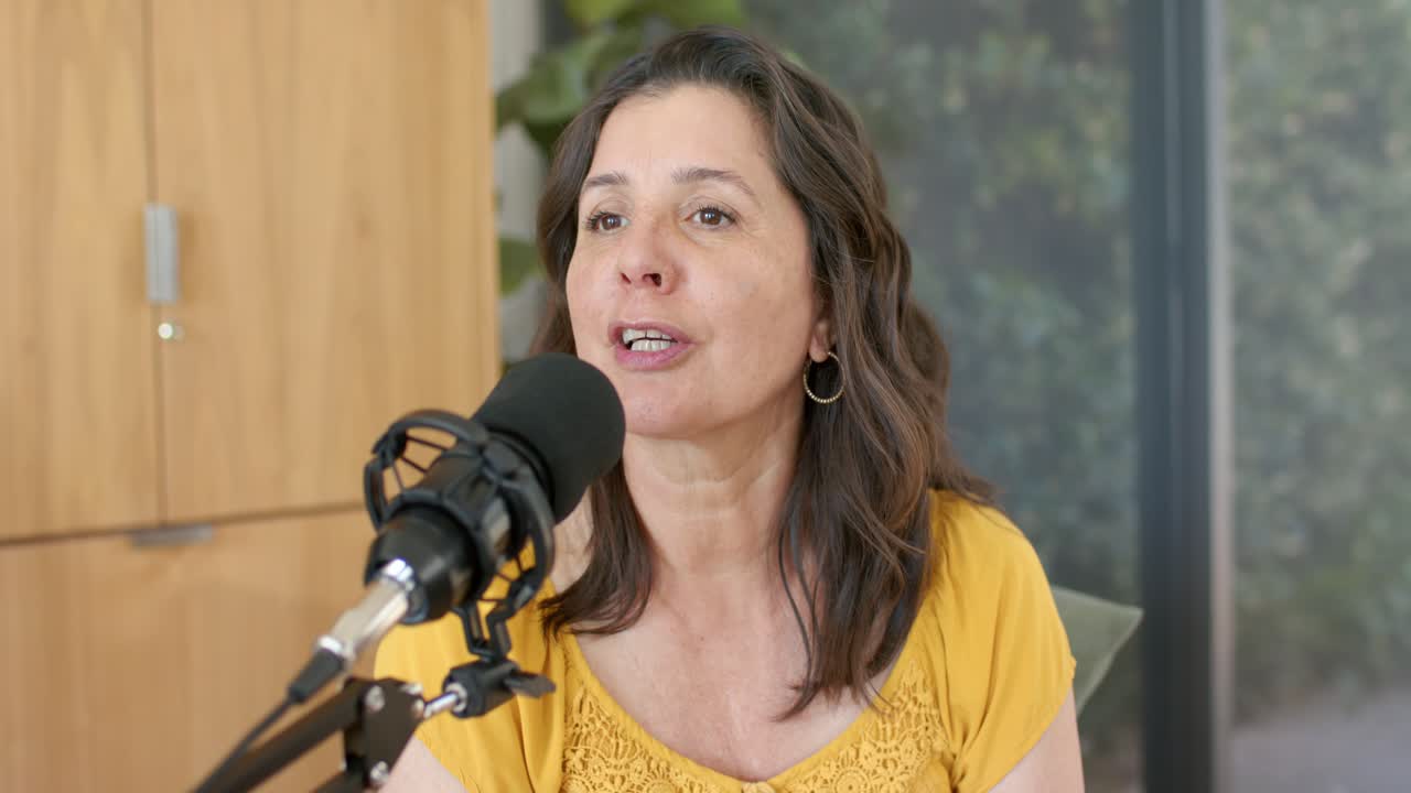 Una donna matura è seduta dietro a un microfono, parla mentre conduce un podcast da casa, indossando un top color senape.