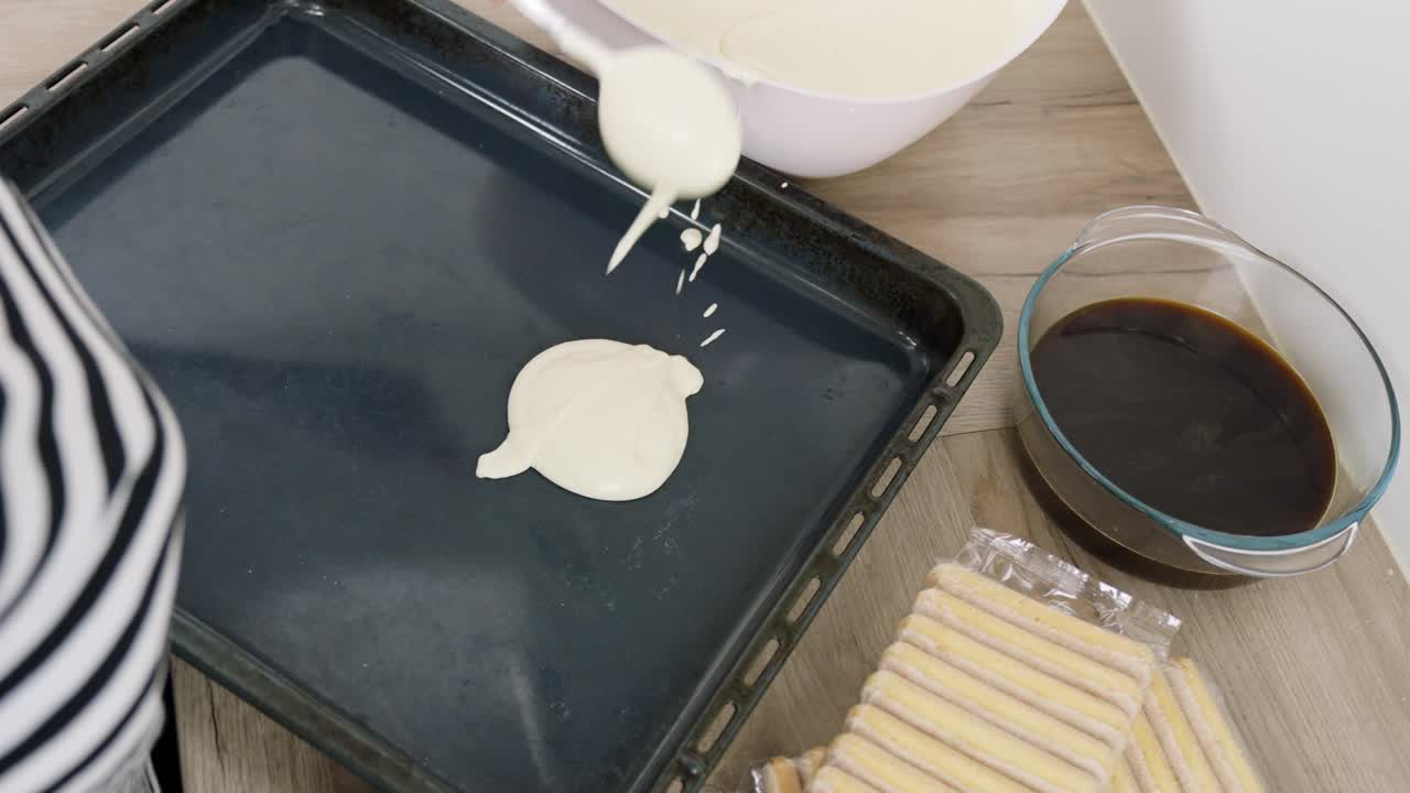Skje sprer kremet mascarponeblanding på et stekebrett for å begynne å sette sammen tiramisu, ved siden av kaffeskål og ladyfingerkjeks, fanget i sanntid ovenfra.