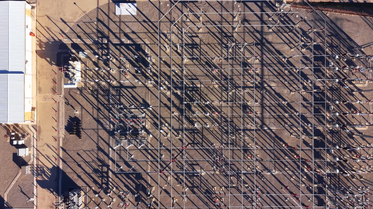 Overhead view of a large electrical substation with metal framework, power lines, transformers, and long shadows cast across a gravel yard beside an industrial building