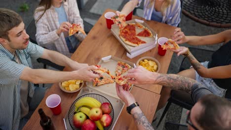 Top-view-of-a-happy-group-of-friends-sorting-out-pizza-from-paper-packaging-during-a-shared-lunch-at-a-table-in-the-courtyard-of-the-house