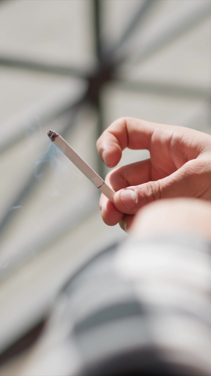 Primo piano di una mano che tiene e muove leggermente una sigaretta spenta con cenere sulla punta. Lo sfondo è leggermente sfocato, suggerendo un ambiente interno o esterno con luce naturale. La mano rimane a fuoco.