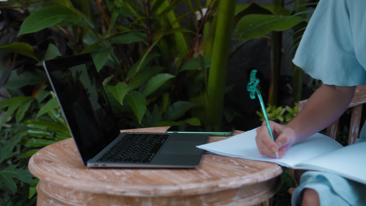 A young European girl in a blue dress remotely online working on laptop and looking into the screen on the backyard with green plants