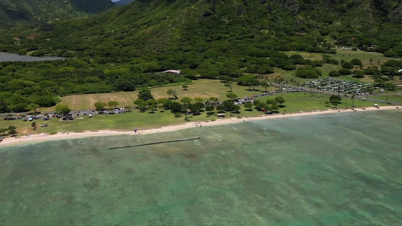 Aerial drone view of a calm turquoise shoreline in Hawaii, showing clear shallow waters and a peaceful sandy beach with greenery beyond. A serene tropical coastal landscape perfect for relaxation