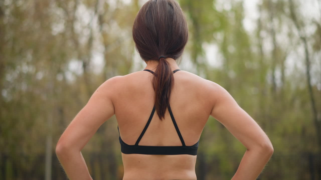 Back view of a lady wearing black singlet and leggings, standing outdoors with hands placed on her waist, showcasing a confident posture, athletic physique, and relaxed demeanor in a natural setting