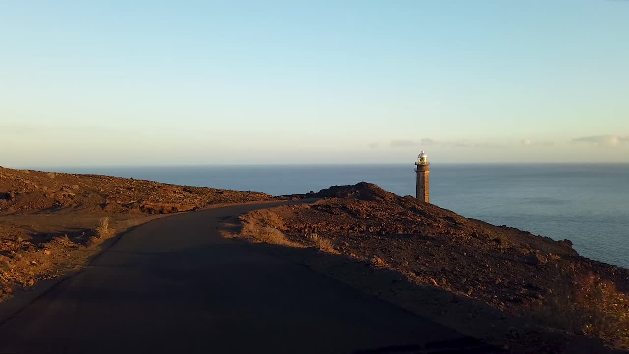 conduciendo por la carretera al atardecer hasta el faro que se eleva sobre la costa rocosa de la isla heirro, faro de la orchilla