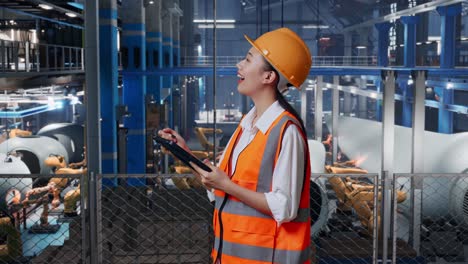 vista lateral de una ingeniera asiática con casco de seguridad de pie en una fábrica de fabricación de turbinas eólicas. tomando nota en la tableta y mirando a su alrededor mientras el brazo robótico trabaja