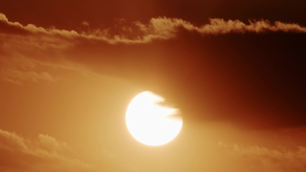 Närbild av en stark sol delvis täckt av tjocka rörliga moln, som glöder i intensiva guldorange solnedgångstoner.