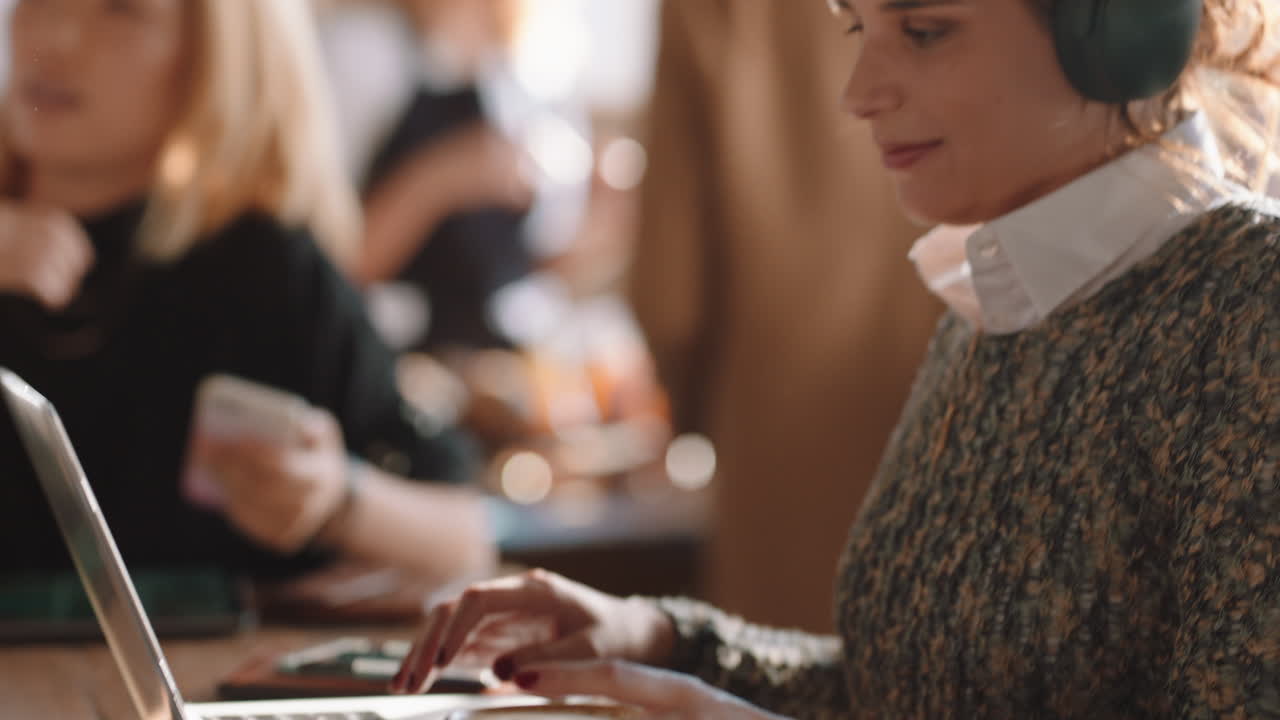 young woman using laptop working in cafe typing email sharing messages on social media wearing headphones enjoying listening to music in busy restaurant drinking coffee