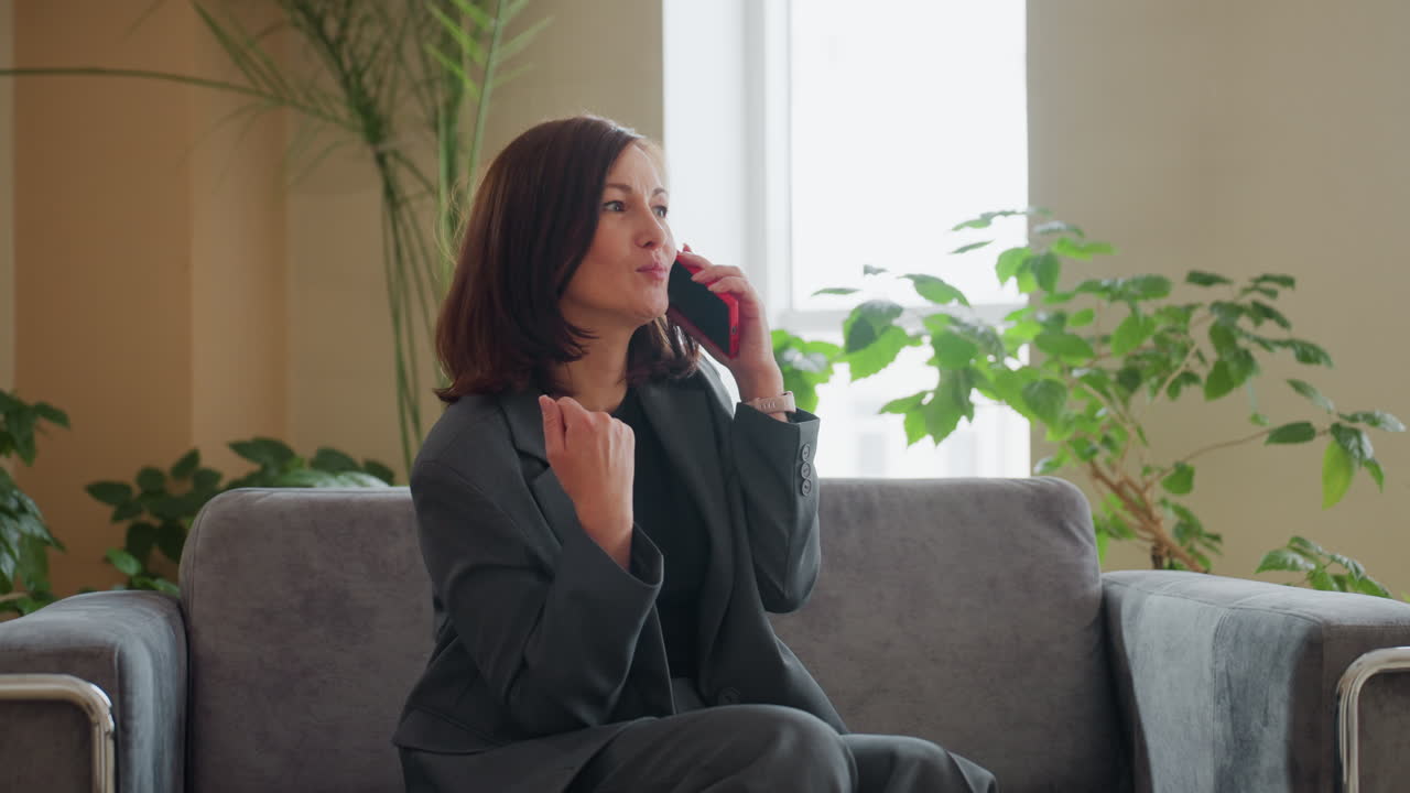 Concerned businesswoman in gray suit sitting on couch speaking on phone with serious look and hand gesture in bright indoor space with houseplants and soft daylight streaming through window