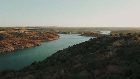 revealing aerial shot of winding long river with bridge behind hills, scenic landscape