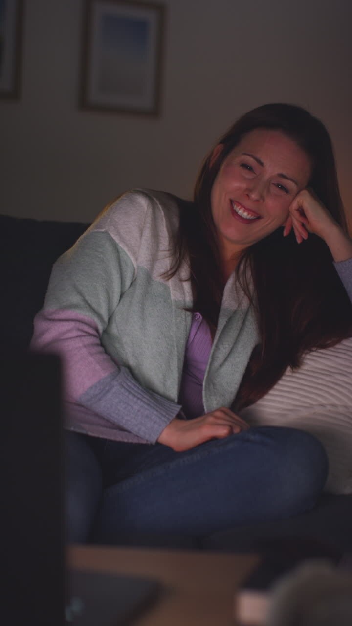 Vertikales Video einer lächelnden Frau, die nachts zu Hause auf einem Sofa sitzt und einen Film oder eine Sendung auf einem Laptop oder Fernseher streamt oder ansieht