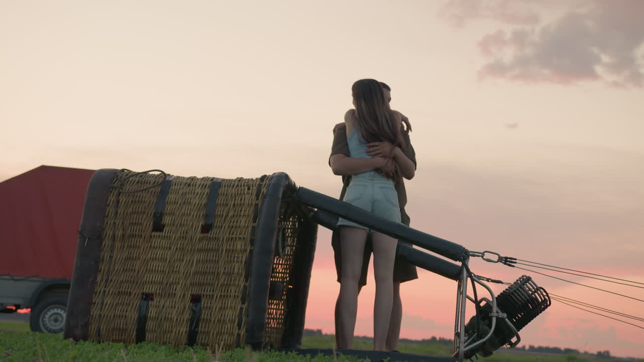 young couple exits wicker hot air balloon basket after landing on green field during colorful sunset with metal burner visible and transport trailer parked nearby under fading light