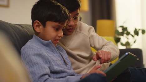 two young boys sitting on sofa at home playing games or streaming onto digital tablet