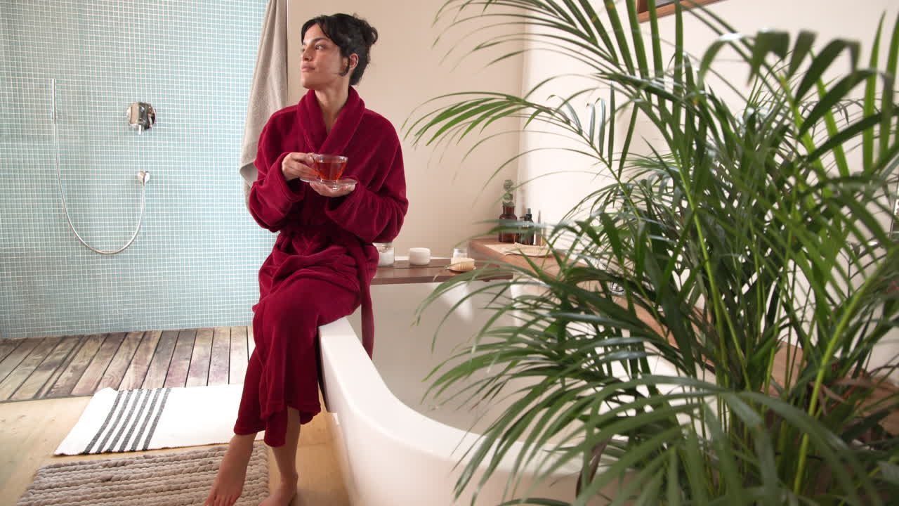 Relaxing at home, woman in red robe enjoying tea by bathtub, copy space
