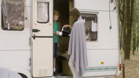 Happy-mother-pouring-tea-from-kettle-into-her-son's-cup-while-standing-by-the-campervan-in-the-forest