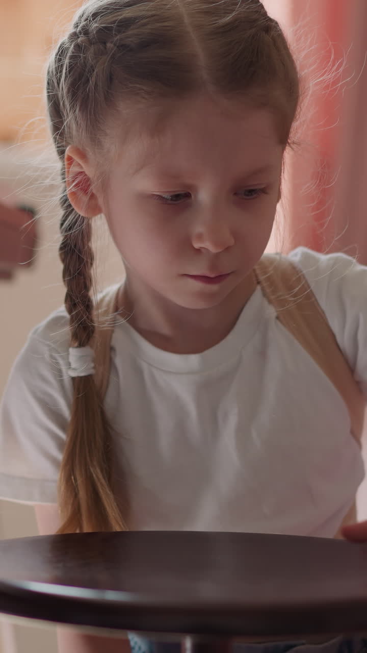 Calm little girl with braided hair spins piano stool by synthesizer piano on floor. Thoughtful child with back braces adjusts chair before music lesson
