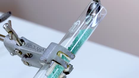 A test tube with green powder secured by a clamp, observed in a laboratory setting.