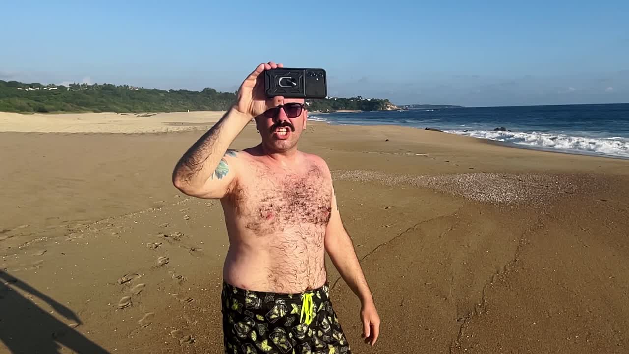 슬로우 모션 백인  ⁇ 한 관광객이 해가 지는 동안 해변에서 걸어서 스마트폰으로 화상 통화 컨퍼런스에서 이야기하고 있습니다.